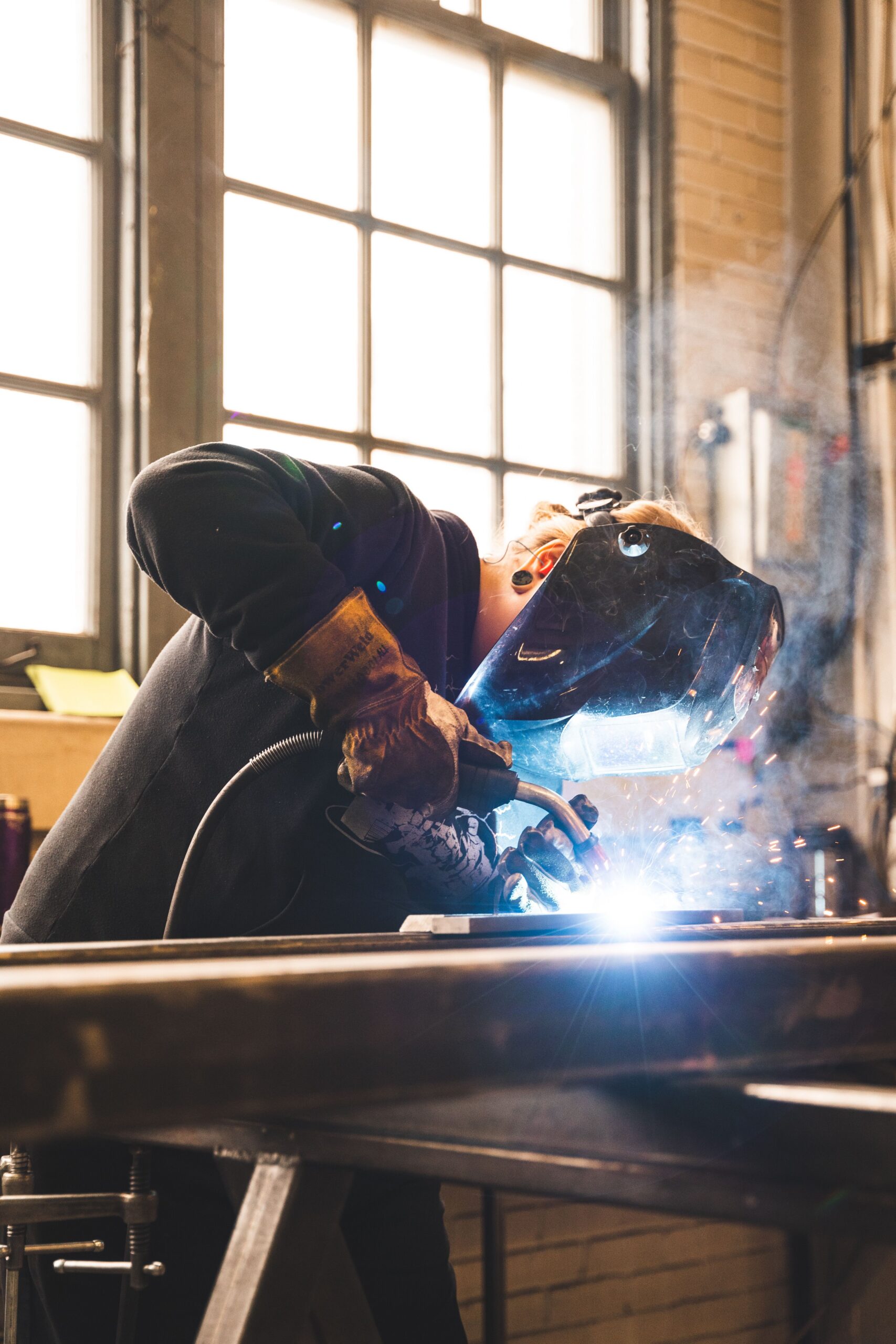 A welder wearing a protective helmet and gloves joins metal in a workshop.